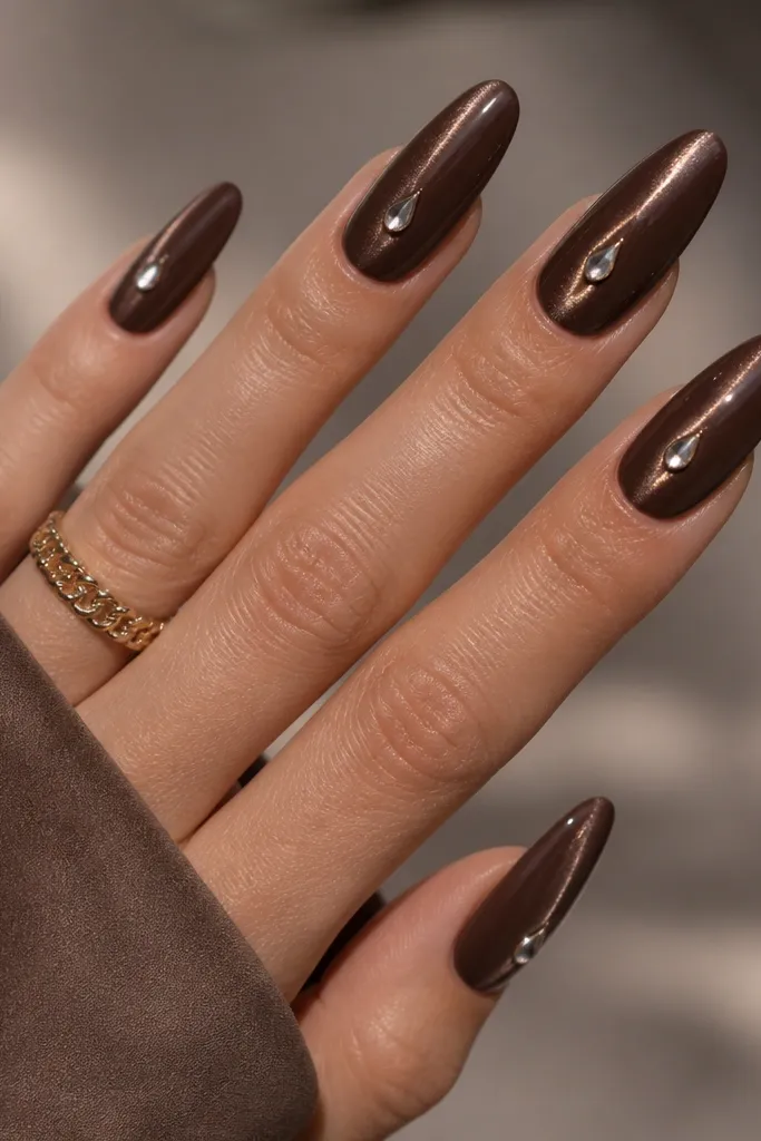 Long almond nails in cocoa brown with a centered cat-eye stripe. On each nail, a single tiny teardrop rhinestone sits slightly above the midline, aligned to the stripe.