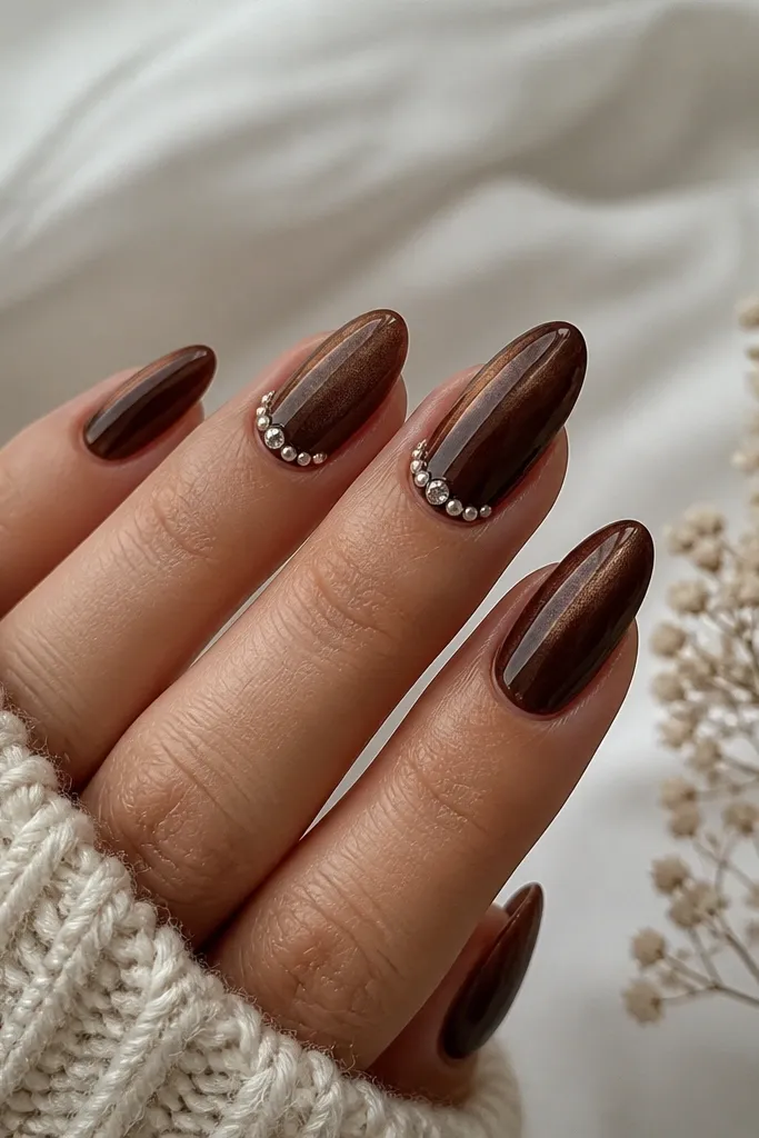 Long almond nails in espresso brown cat eye with a centered stripe. Along the cuticle line on two accent nails, there is a thin row of tiny pearls mixed with one or two clear crystals.