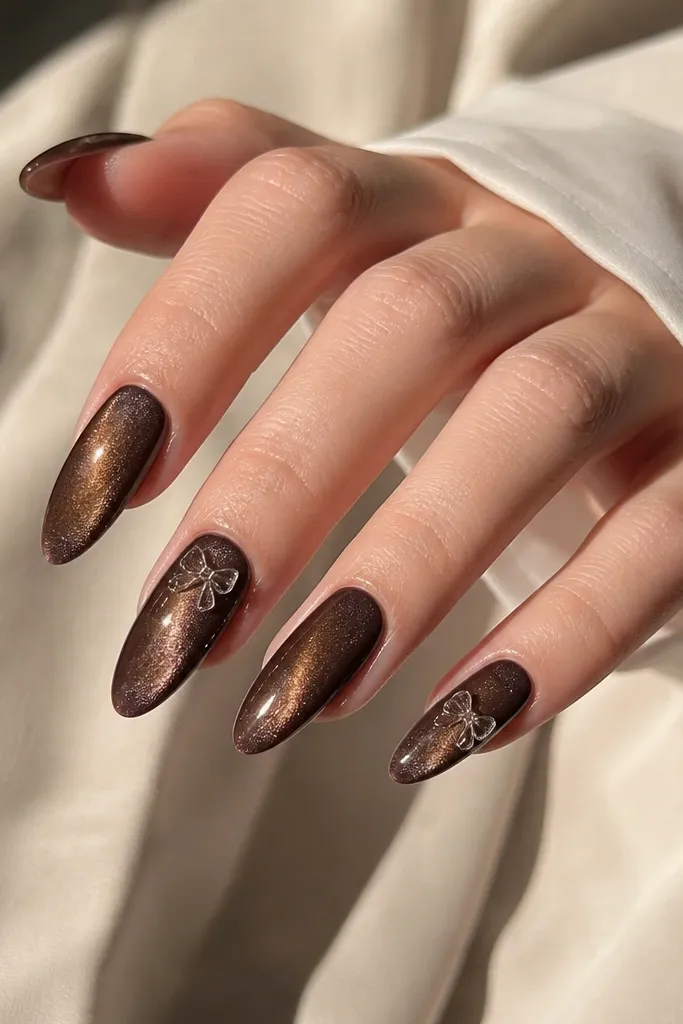 Long almond nails with espresso brown cat-eye stripe; one or two nails have a small clear 3D bow charm near the cuticle area.