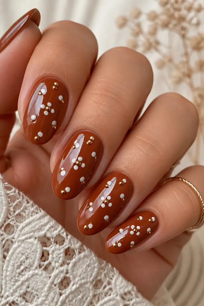 A rust brown base with alternating white polka dots and tiny gold studs near the dots. Each nail has a neat cluster around the center area. Glossy top coat.