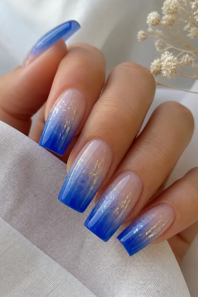 Long square nails with cobalt blue at the tips fading into clear at the cuticle; thin aurora chrome flakes sit in the gradient like scattered light.