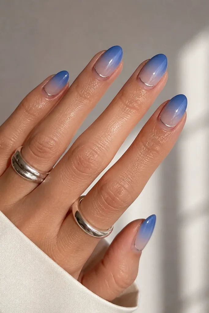 Medium almond nails with cornflower blue fading into sheer at the cuticle; a thin silver chrome band hugs the cuticle line like a halo.