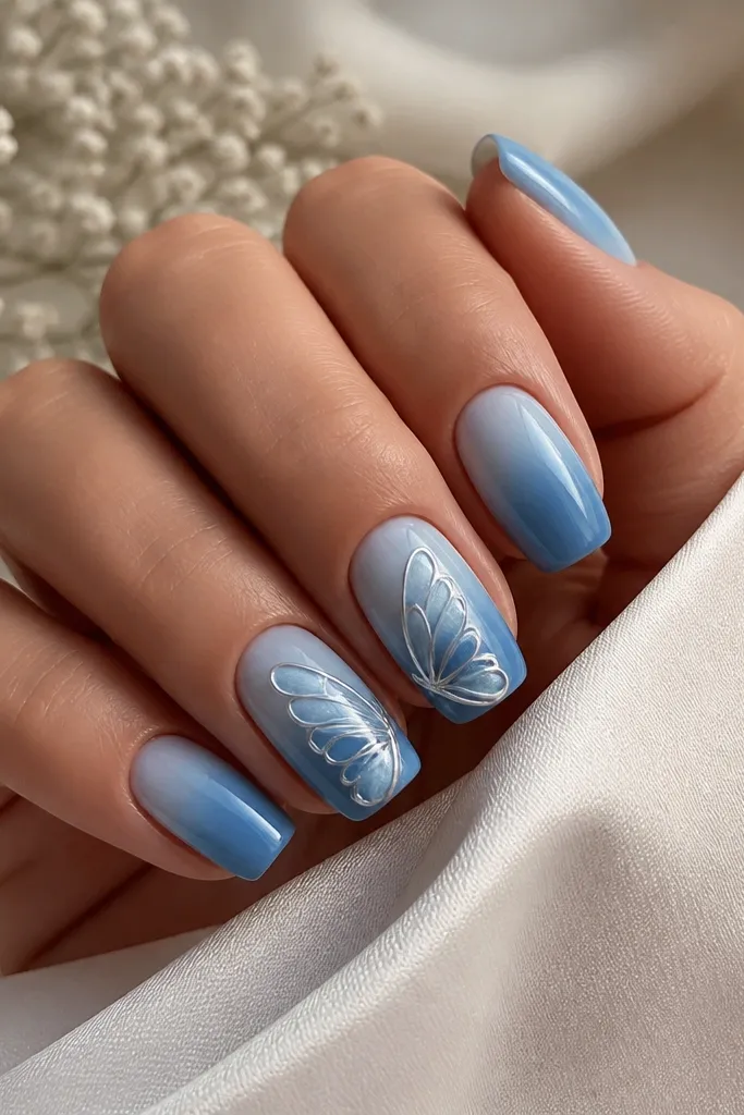 Medium squoval nails with blue ombre; on two accent nails, chrome butterfly wing shapes sit diagonally, mirrored in pale blue-silver.