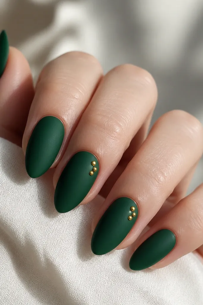Short stiletto nails painted deep emerald green matte; two nails have a tiny cluster of three gold studs near the cuticle on one side; other nails are plain matte.