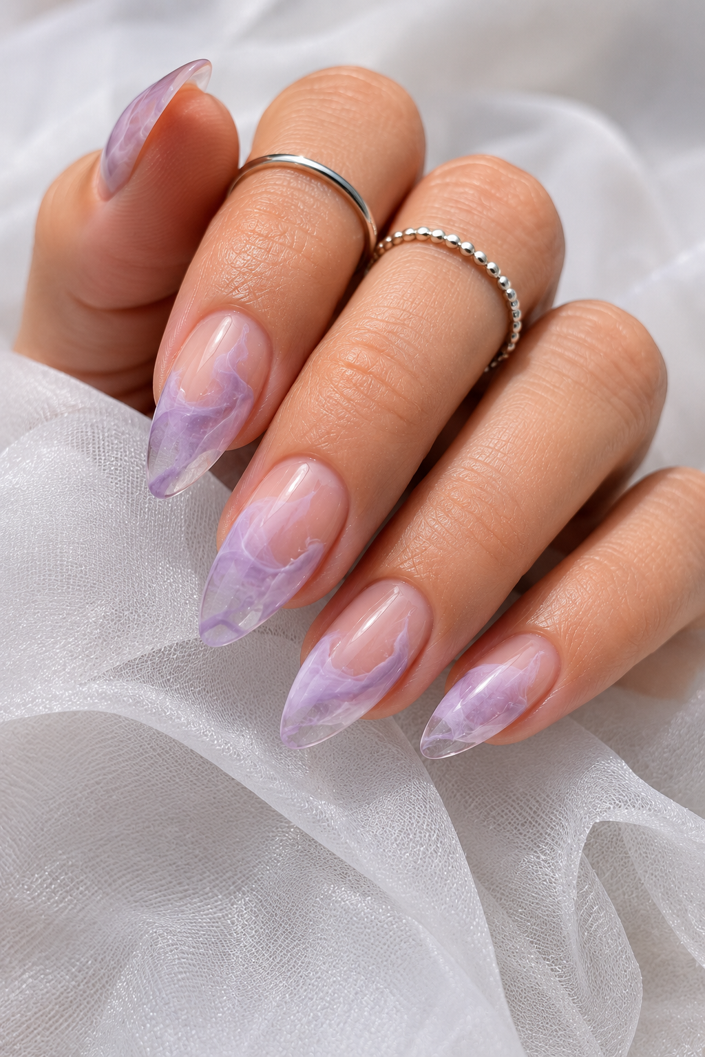 Short stiletto nails with a glossy clear-to-lilac gradient; on top, soft lilac swirls resembling small clouds across the center; background looks shiny and jelly-like.