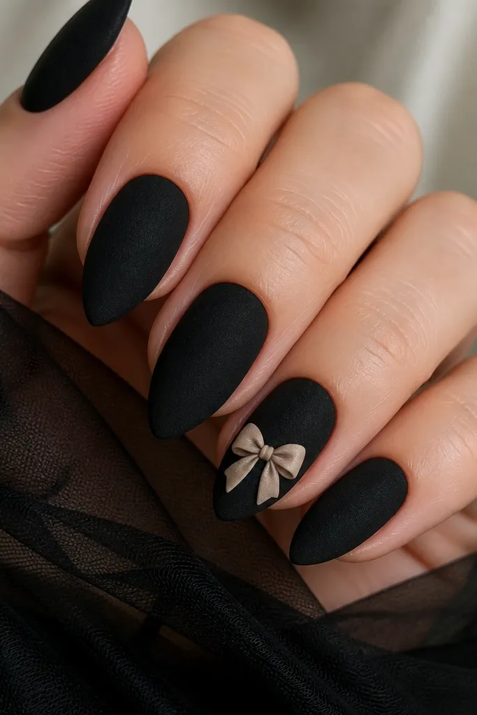 Short stiletto nails in black velvet matte finish; on one accent nail there is a nude bow with two loops and a small center knot near the tip area; matte texture visible.