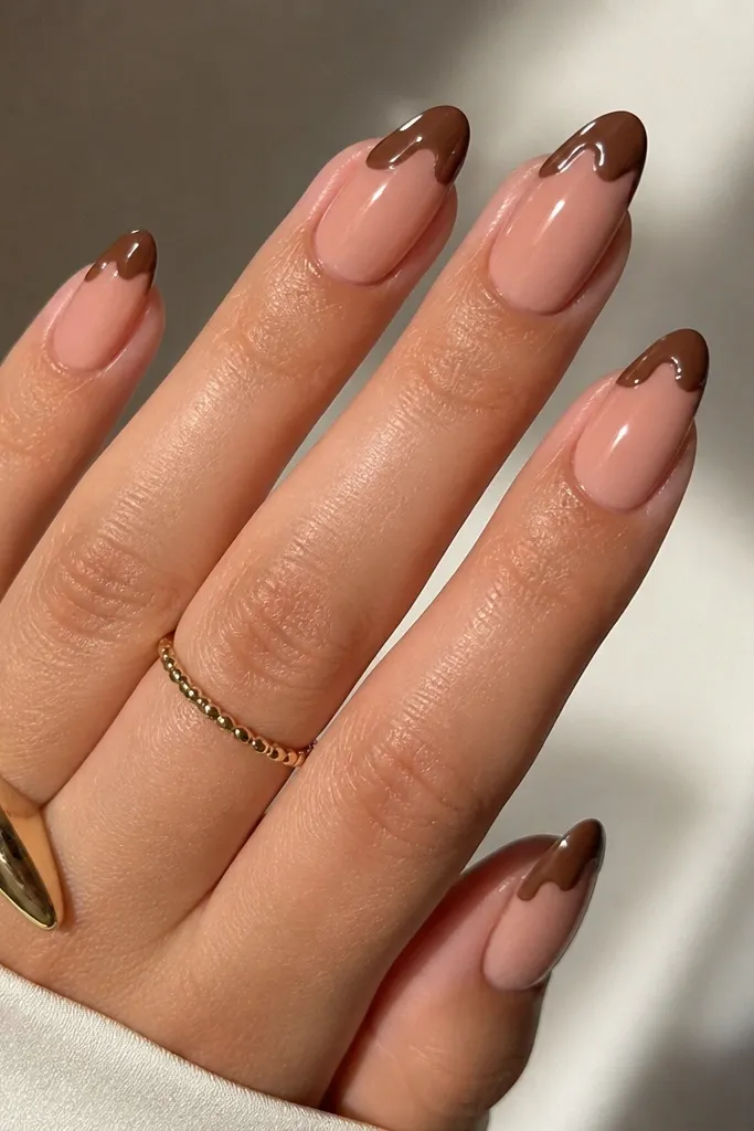 Medium almond nails with a nude base. The French tip is chestnut brown, but the tip edge has a small swirl pattern instead of a straight line. Glossy finish.