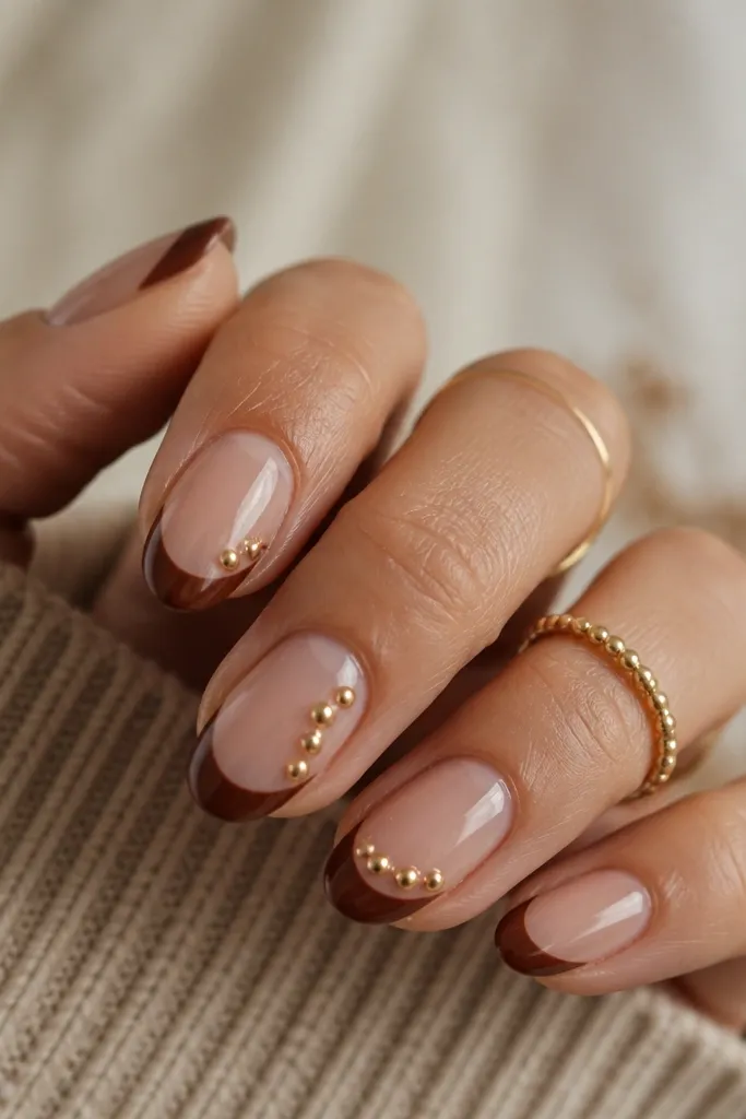 Short squoval nails with a chestnut French tip. On each tip there are one or two tiny gold studs placed near the side edges, like small accents. Glossy finish.