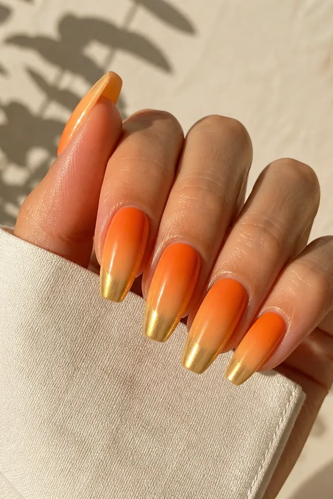 Medium coffin nails with a tangerine-to-peach ombre that fades into bright gold chrome at the tips, reflecting warm sunlight.