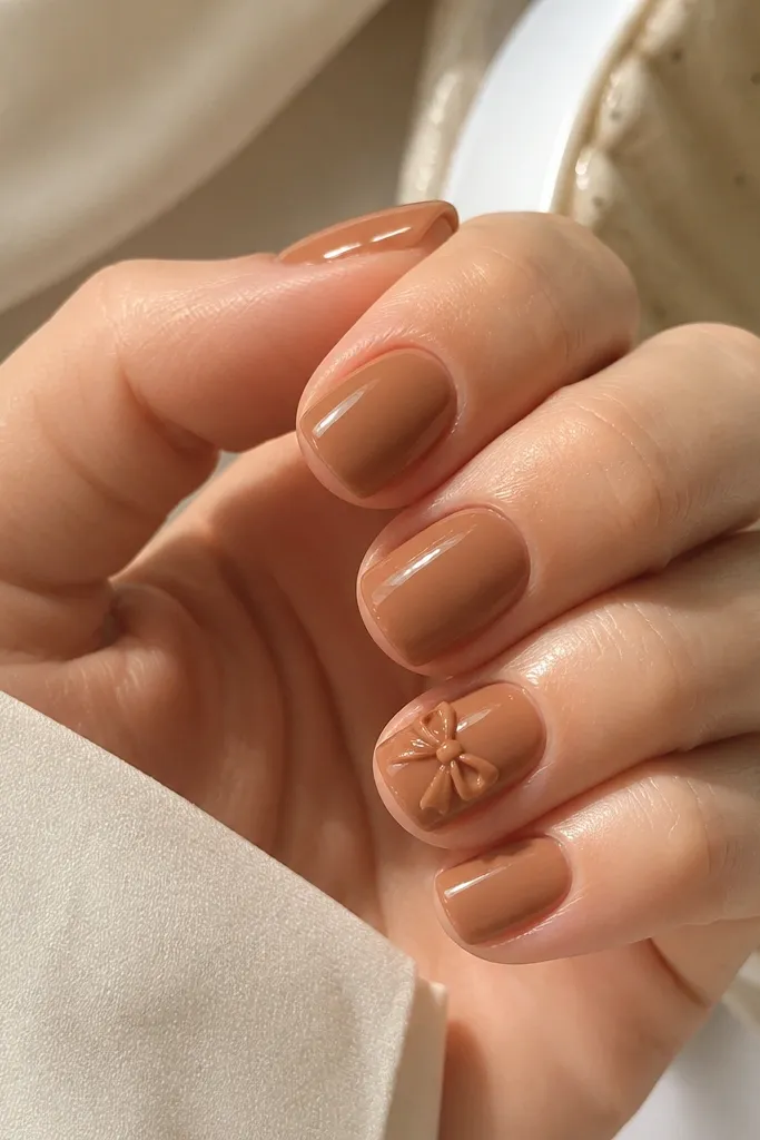 Short squoval nails in caramel nude, with one or two nails featuring a small 3D caramel bow made from gel or nail art striping, glossy topcoat around it.