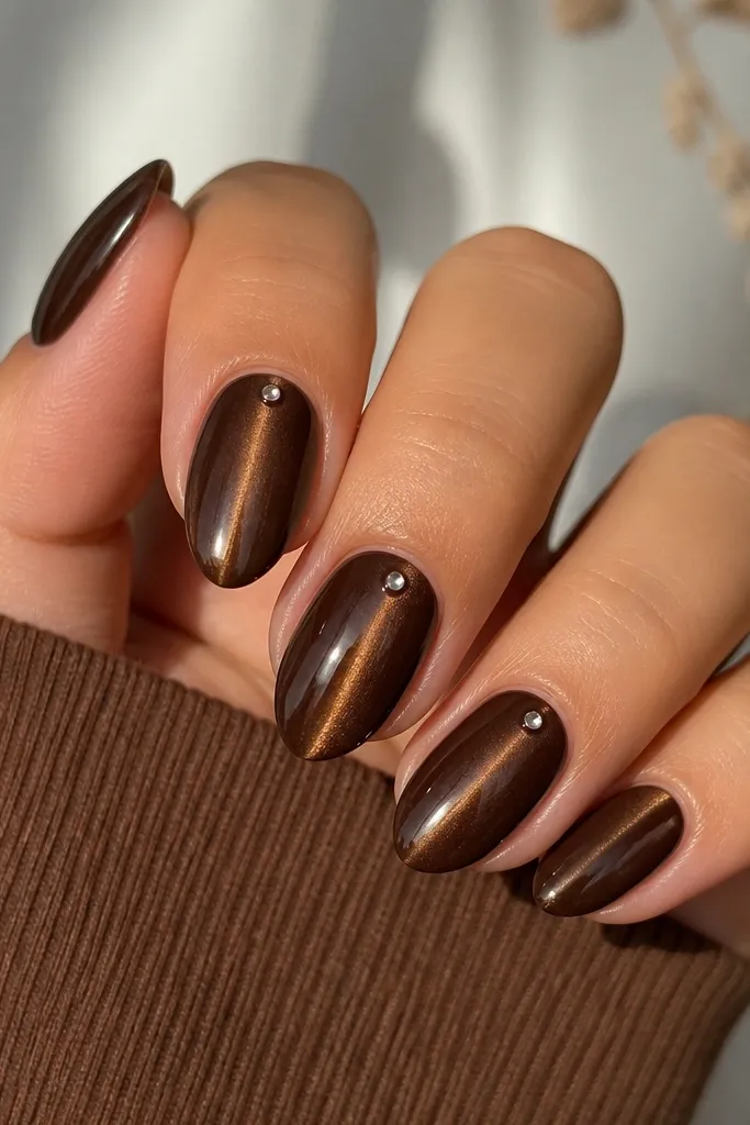 Chocolate brown nails with a centered gold cat-eye streak. On one nail (or two), there's a single tiny clear rhinestone placed near the cuticle on the streak line. Glossy finish.