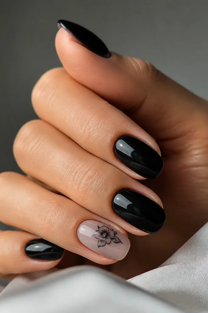 Mostly solid black nails; one accent nail has a small black-and-gray floral sticker placed near the cuticle, sealed under a clear glossy layer.