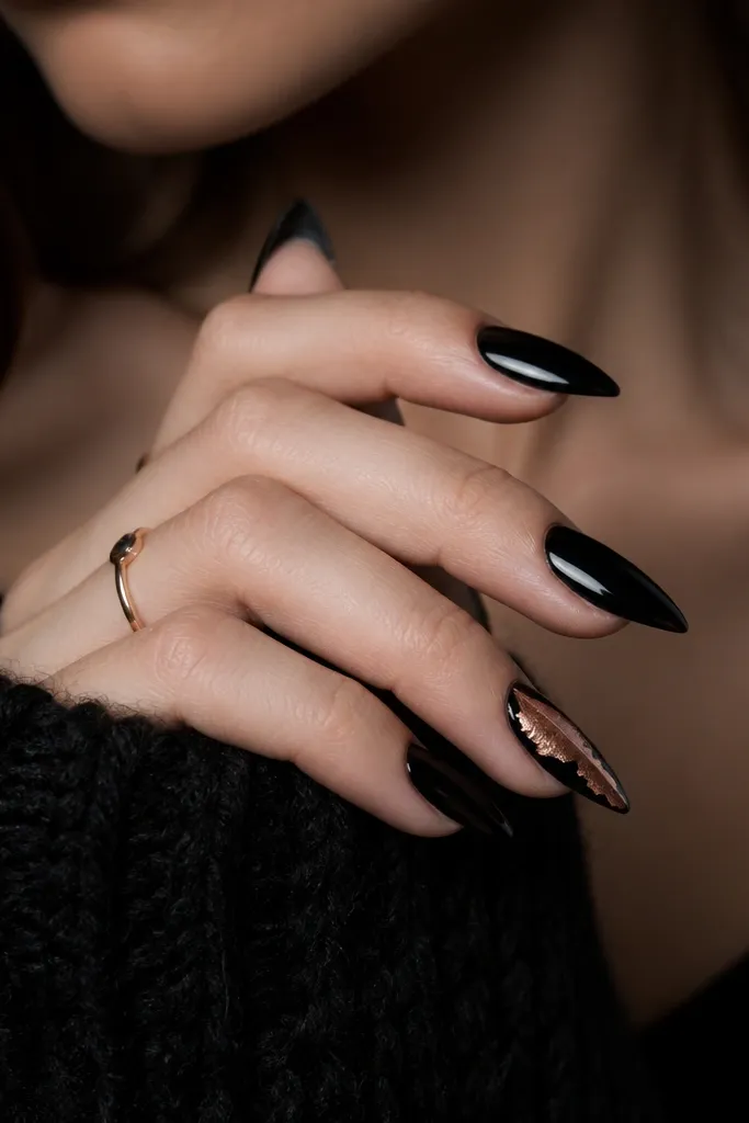Short stiletto nails with solid black; one accent nail has rose gold foil pieces arranged like a diagonal spear from mid-nail to tip.