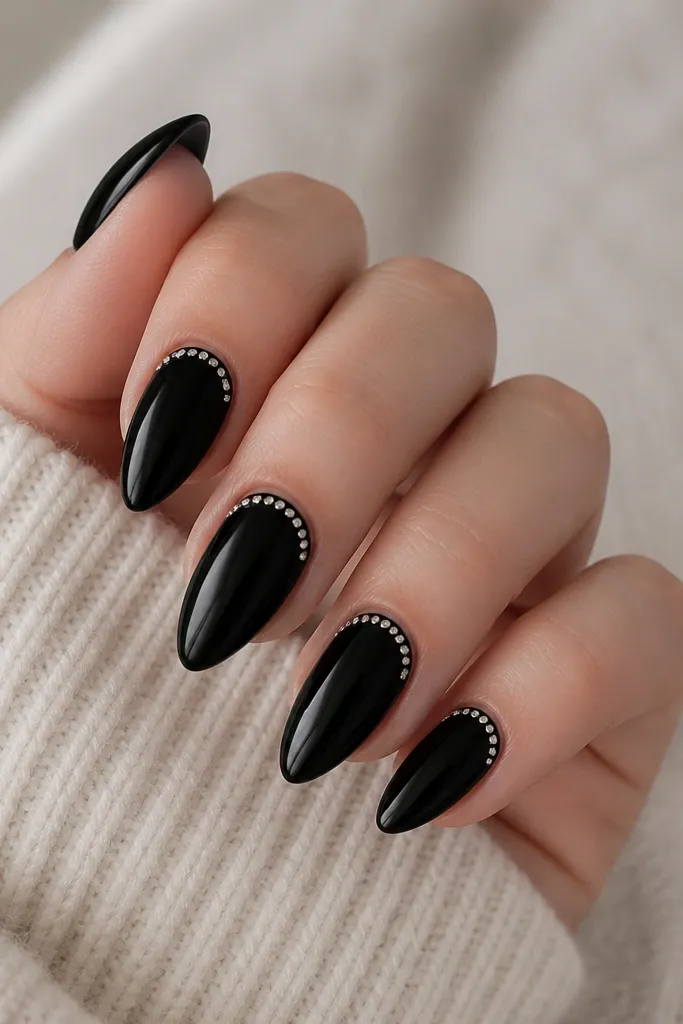 Short stiletto nails with solid black; tiny clear or silver studs line a curved arc just below the cuticle on each nail, all glossy.