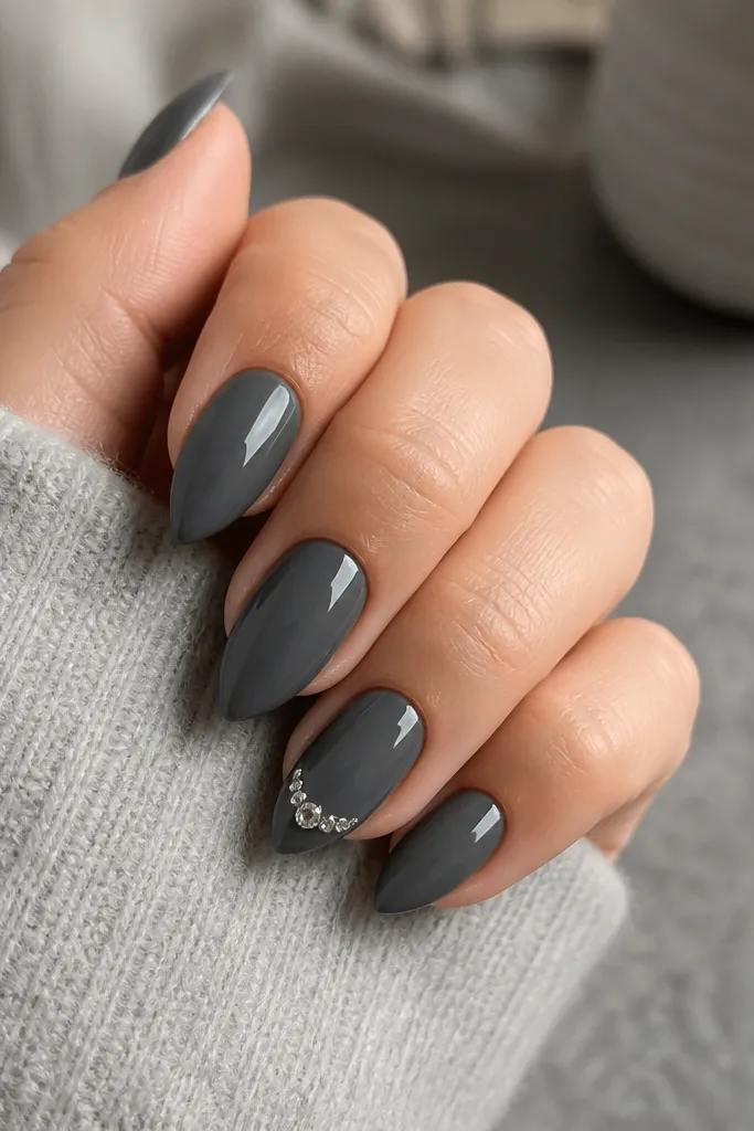 Compact short stiletto nails in glossy charcoal gray. One accent nail has a tiny cluster of clear crystals near the tip, arranged like a small arc.