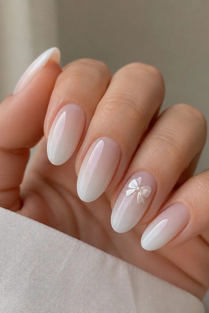 Almond nails with a milky white ombre fading to clear. On one accent nail, a small bow tie shape is made from pearl chrome near the cuticle center. Other nails keep the ombre simple with a subtle shine.