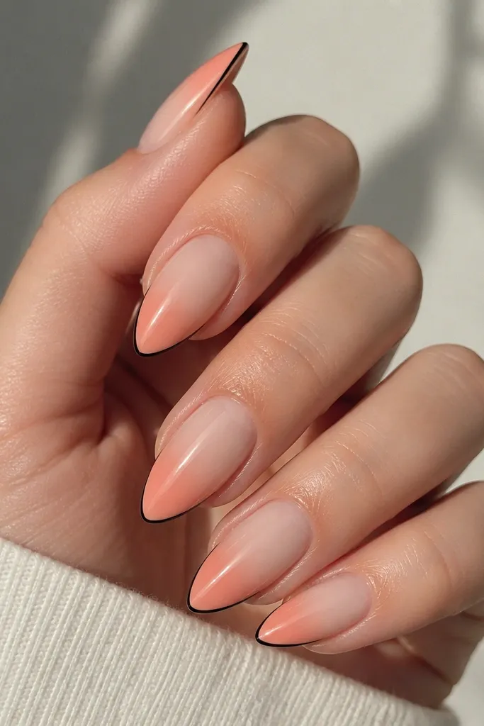 Short stiletto nails with a soft peach ombre that fades from nude near the cuticle to peach at the tip. A thin black line traces the outer edge of the peach tip like eyeliner.