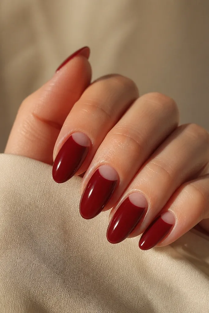 Deep cherry red nails with a neat negative-space half-moon near the cuticle on two accent nails per hand; the rest are solid cherry gloss.