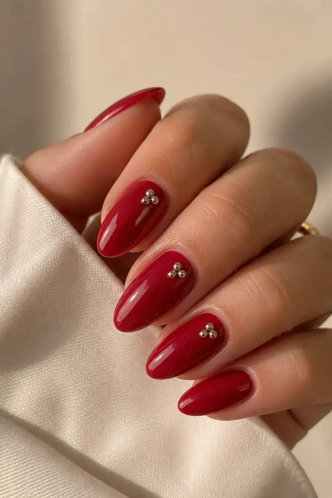 Deep cherry red glossy nails with one nail on each hand featuring a tiny silver chrome dot near the center, like a pinpoint highlight.