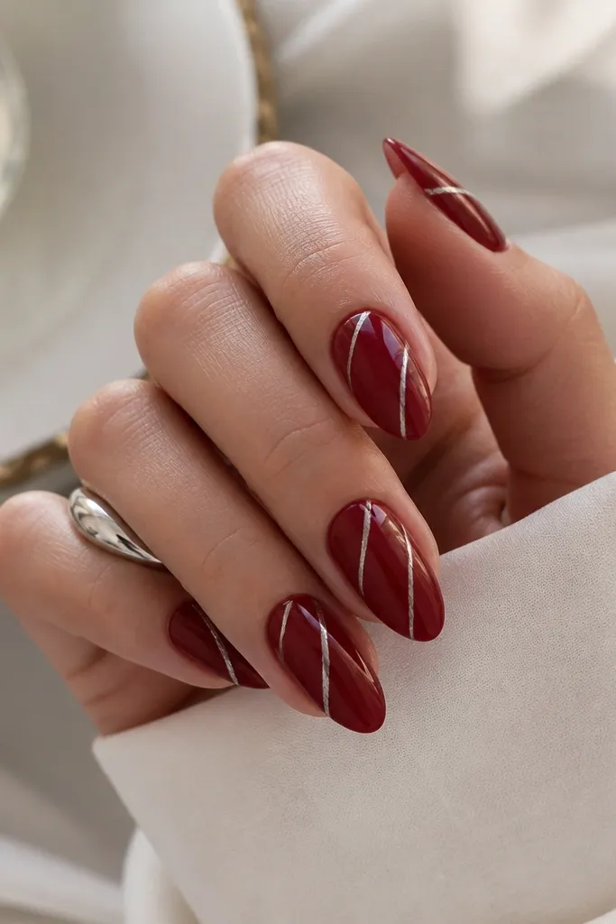 Medium almond nails painted deep red with thin silver chrome vein lines running diagonally across the nails. The chrome catches light like foil.