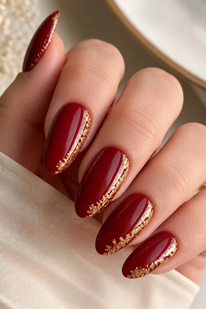 Almond nails painted deep red with thin gold foil leaf pieces concentrated along one side edge of each nail, like a gilded border. Glossy finish.