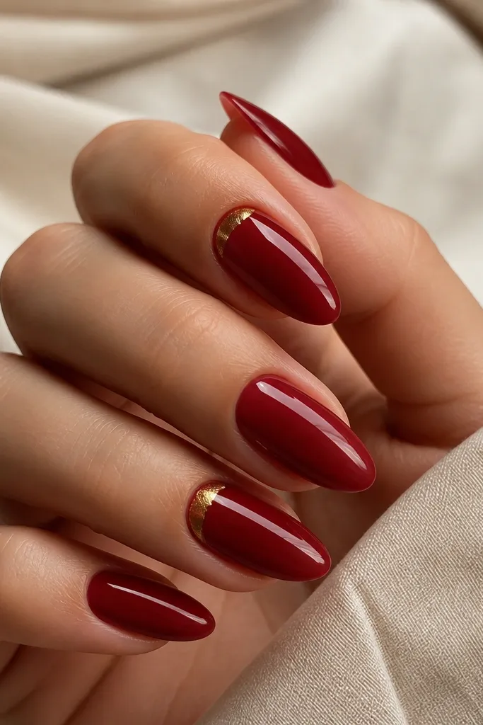 Some almond nails are solid deep red glossy; accent nails have a tiny gold foil crescent near the cuticle, like a small metallic smile.