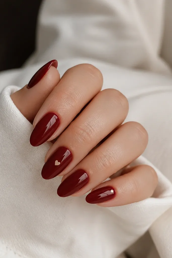 Almond nails are deep red glossy. One accent nail has a tiny cream heart dot near the center, and another has a single cream dot under the cuticle.
