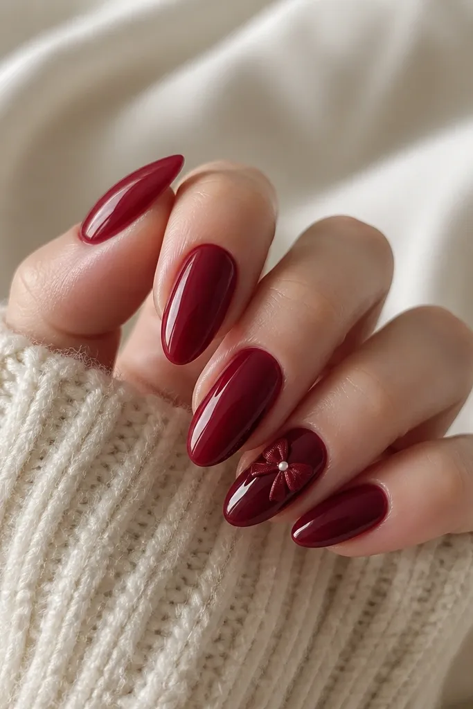 Deep red glossy almond nails with one accent nail featuring a small satin ribbon bow in dark red and a tiny highlight on the bow loops.