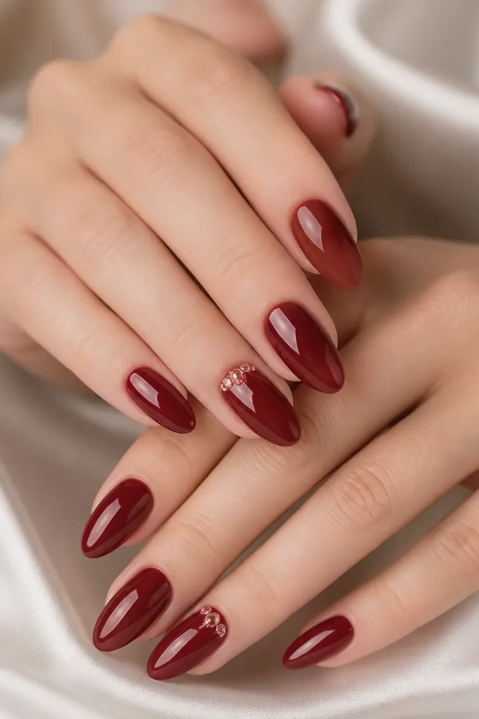 Deep red glossy almond nails with a few tiny rose-pink crystal studs clustered near the cuticle on two accent nails; the crystals catch light.