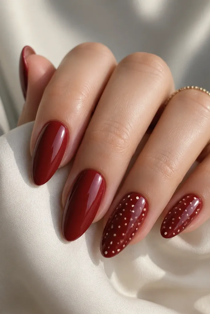 Deep red glossy almond nails with scattered tiny cream dots on two accent nails; dots vary in size but stay small and evenly spaced.