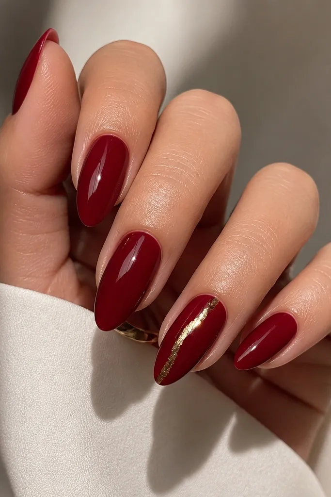 Almond nails in glossy deep red, with one accent nail featuring a thin gold foil strip running diagonally from near the cuticle toward the side of the tip. The gold catches light sharply.