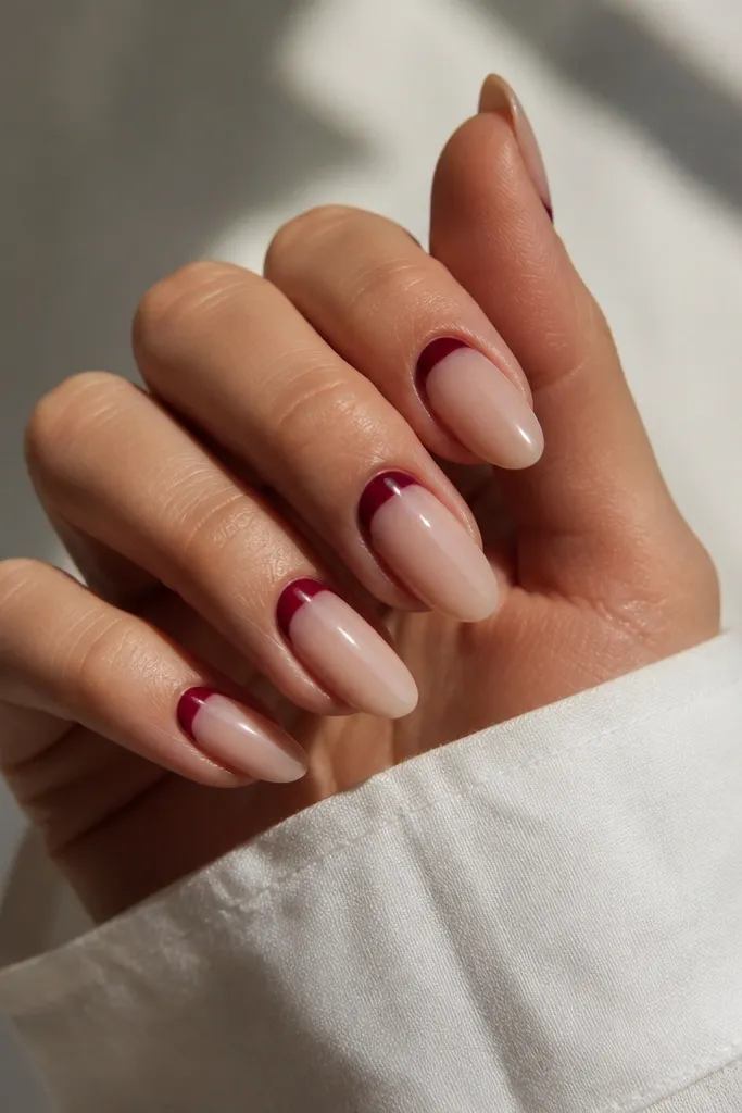 Almond nails with a sheer nude base. Deep red half-moon shapes sit right at the cuticle line, leaving the rest of the nail nude. The nails are glossy.