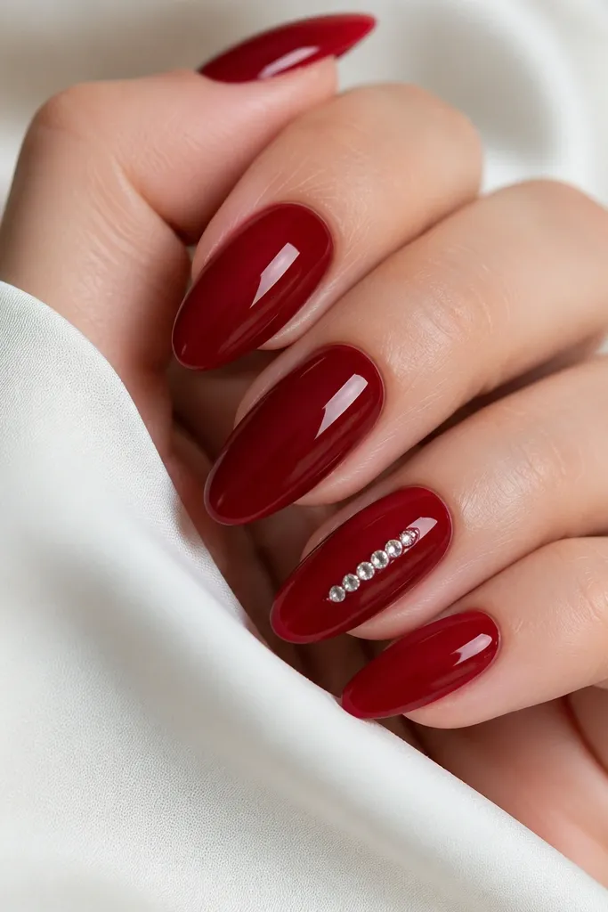 Glossy deep red almond nails with one accent nail featuring a cluster of small clear rhinestones near the center-right of the nail. The stones are aligned and sealed under topcoat.
