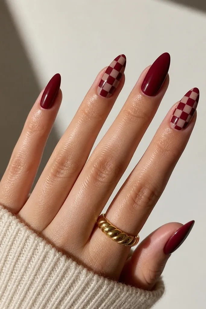 Almond nails in deep red with two accent nails that have a checker pattern using small squares of sheer nude and deep red. The pattern is neat with sharp edges and a glossy topcoat.