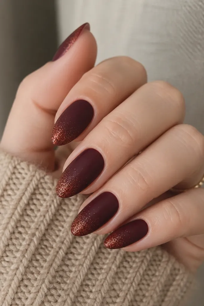 Medium almond nails in deep burgundy. The tips fade into a fine, coppery micro-glitter that looks like glowing embers. Only the outer third has sparkle; the rest stays matte-satin.
