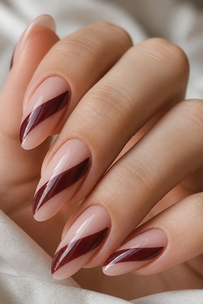 Almond nails with a nude base (sheer pink). A diagonal ribbon crosses from lower left to upper right in glossy deep burgundy. The ribbon has sharp edges and sits on a mostly clear nail.