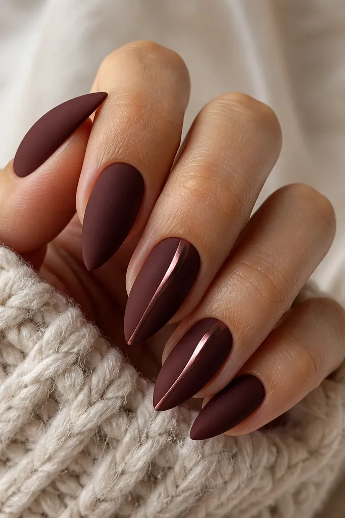 Short stiletto nails in matte burgundy. On two accent nails, a narrow wine-chrome swoosh runs vertically from near the cuticle to the tip, glowing with a soft rose-gold sheen.