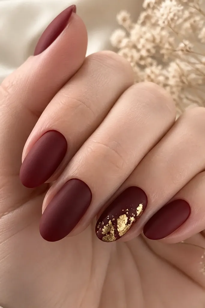 Mostly matte oxblood nails with one accent nail. The accent nail has scattered irregular gold leaf flecks concentrated near the center and lower half, with some flecks catching light.