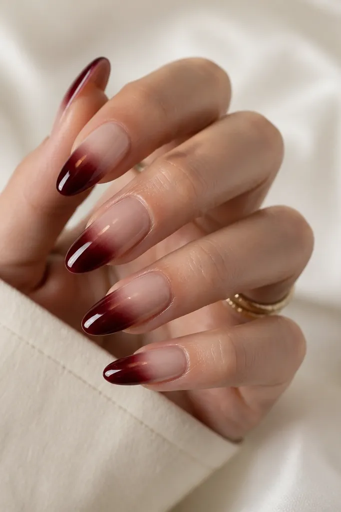 Almond nails. The cuticle area is sheer nude, fading into deep burgundy toward the tip. The finish is glossy and clean, with a soft blur at the blend.