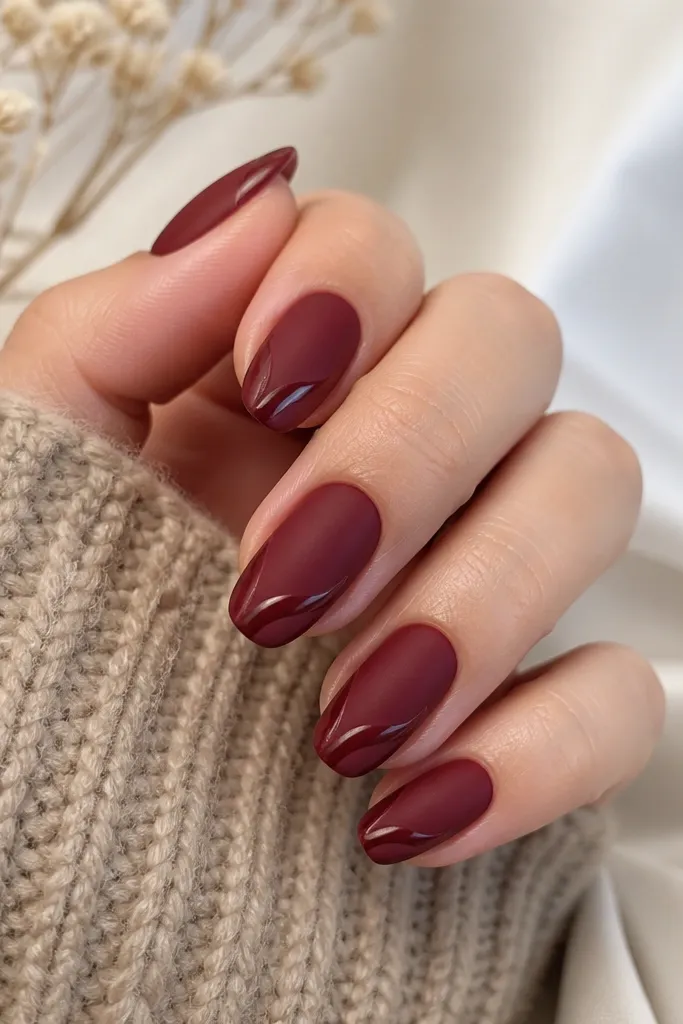 Rounded medium nails. The base is matte burgundy. On each nail, the tip has a glossy swirl line that curves like a leaf stem, staying within the outer third.