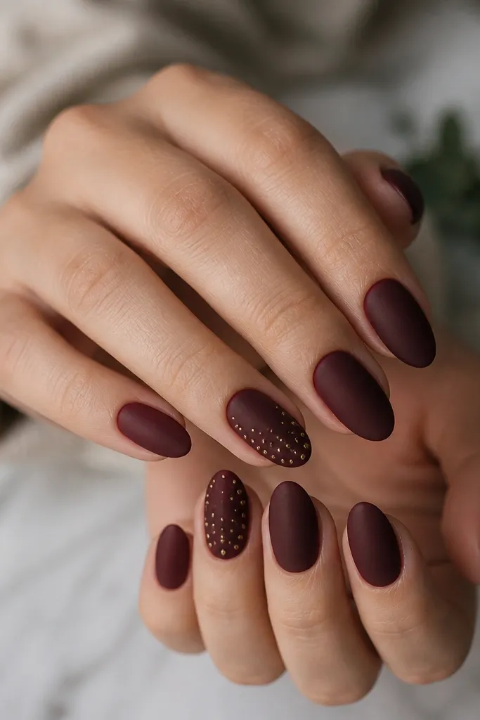 Short almond nails in matte deep burgundy. Two accent nails have tiny flat micro-studs arranged in a diagonal gradient, with more studs near the tip and fewer near the middle.