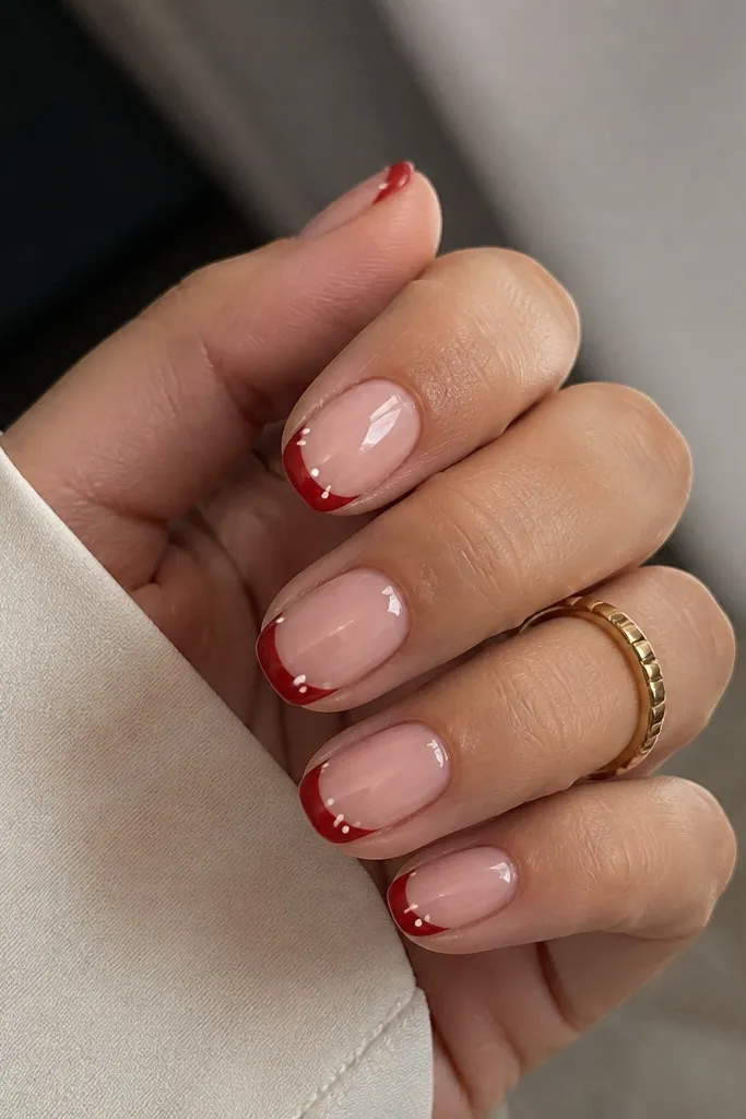 Short nails with a nude base. The French tips are deep red, but the tip edge is curved like a swirl. In the corners near each side of the tip, there are two tiny white dots.