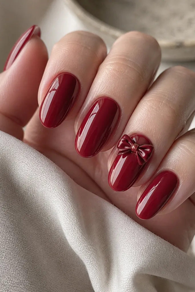 Glossy deep cherry nails. On one thumb nail, a 3D ribbon bow sits near the cuticle: deep cherry bow with a tiny gold center knot. Other nails are solid red.