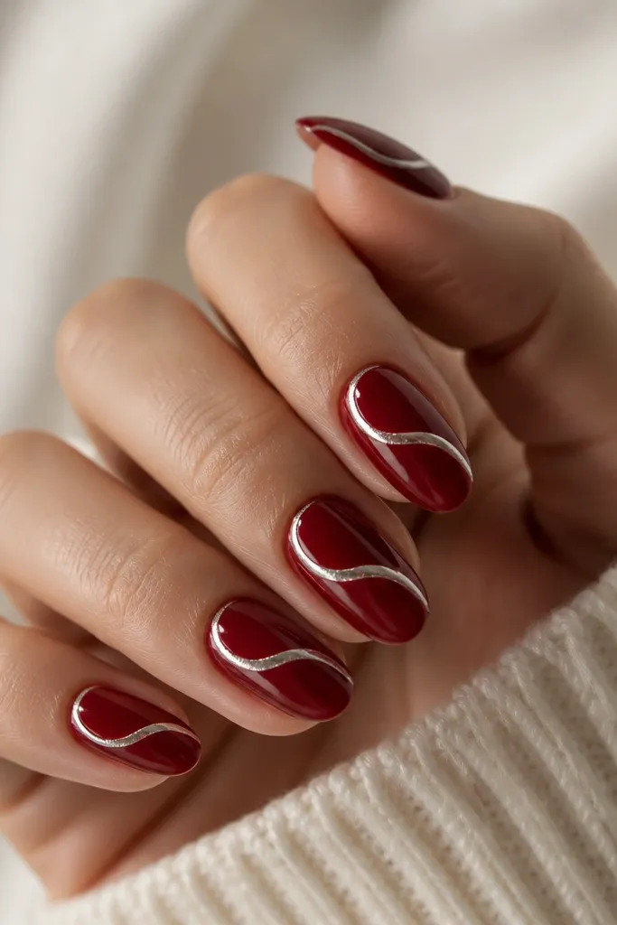 Short almond nails with deep red base; a single chrome swirl ribbon wraps from the side near the cuticle to the center, then curves toward the tip.