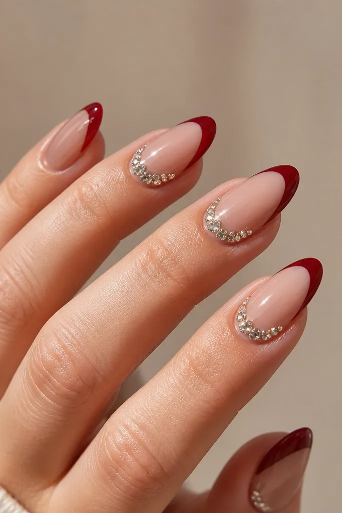 Long almond nails with a glossy nude base; deep red french tips and a small cluster of clear rhinestones at the outer corner of each tip.