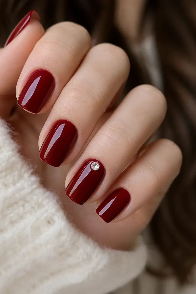 Glossy deep red nails with one accent nail featuring a single small clear rhinestone placed near the cuticle center; the stone reflects light like a bead.