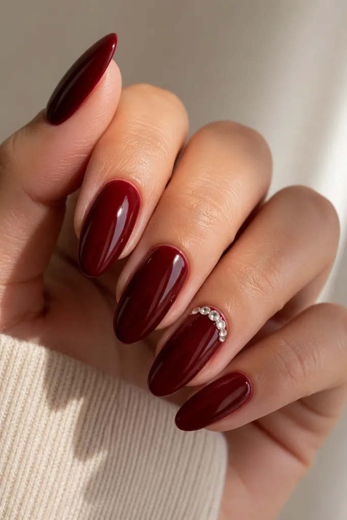 Almond nails in deep red with a small halo of rhinestones around the cuticle on one accent nail; the rest are plain glossy deep red.
