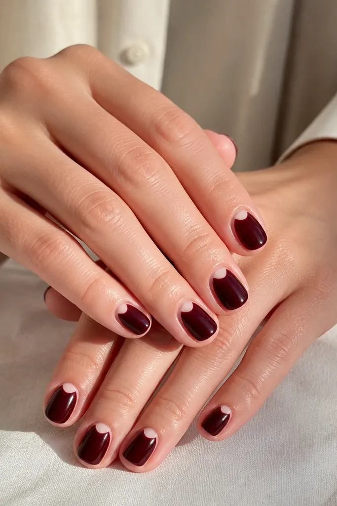 Short nails in deep black-cherry red with a small negative-space half-moon near the cuticle, clean and symmetrical on each nail.