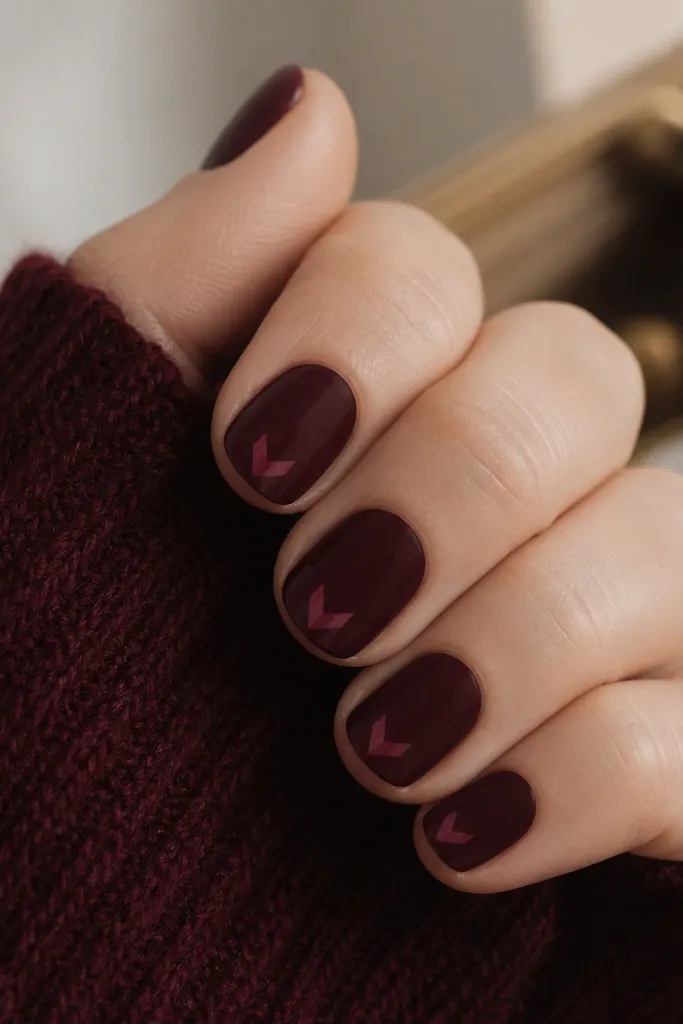 Short nails painted oxblood with a tiny chevron pattern in a slightly lighter burgundy centered near the tip.
