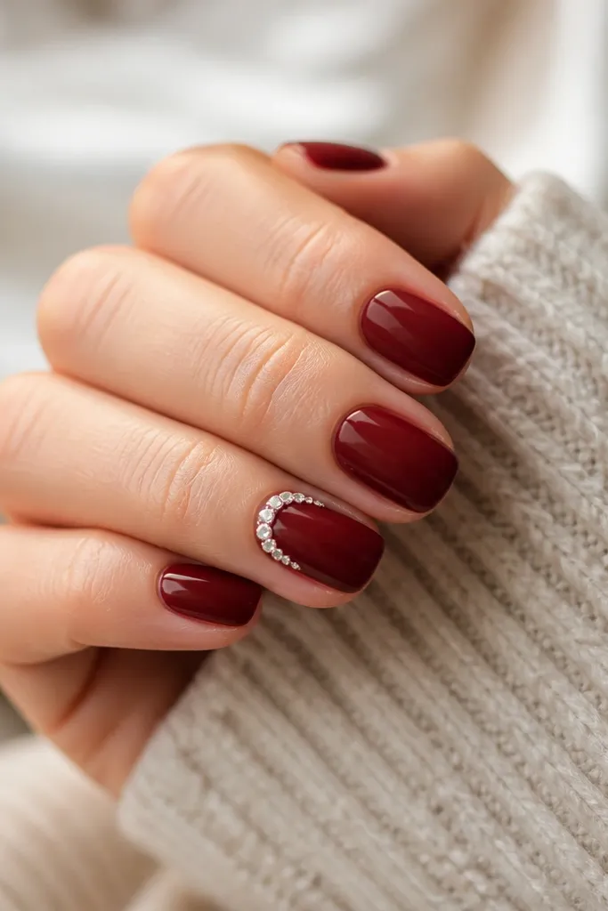 Short deep red nails with one accent nail featuring a small semicircle of tiny clear rhinestones around the cuticle area.